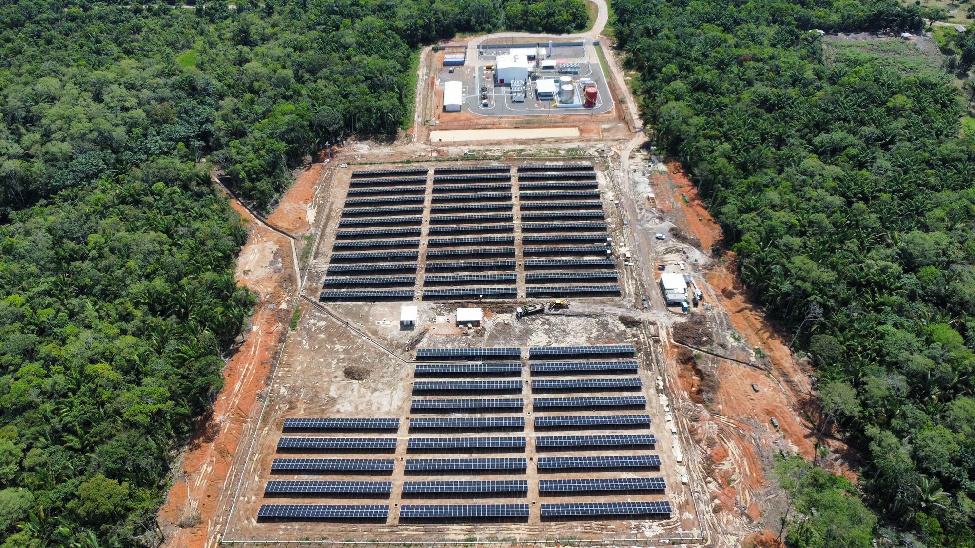 Vista aérea de usina solar cercada de floresta, com subestação ao fundo
